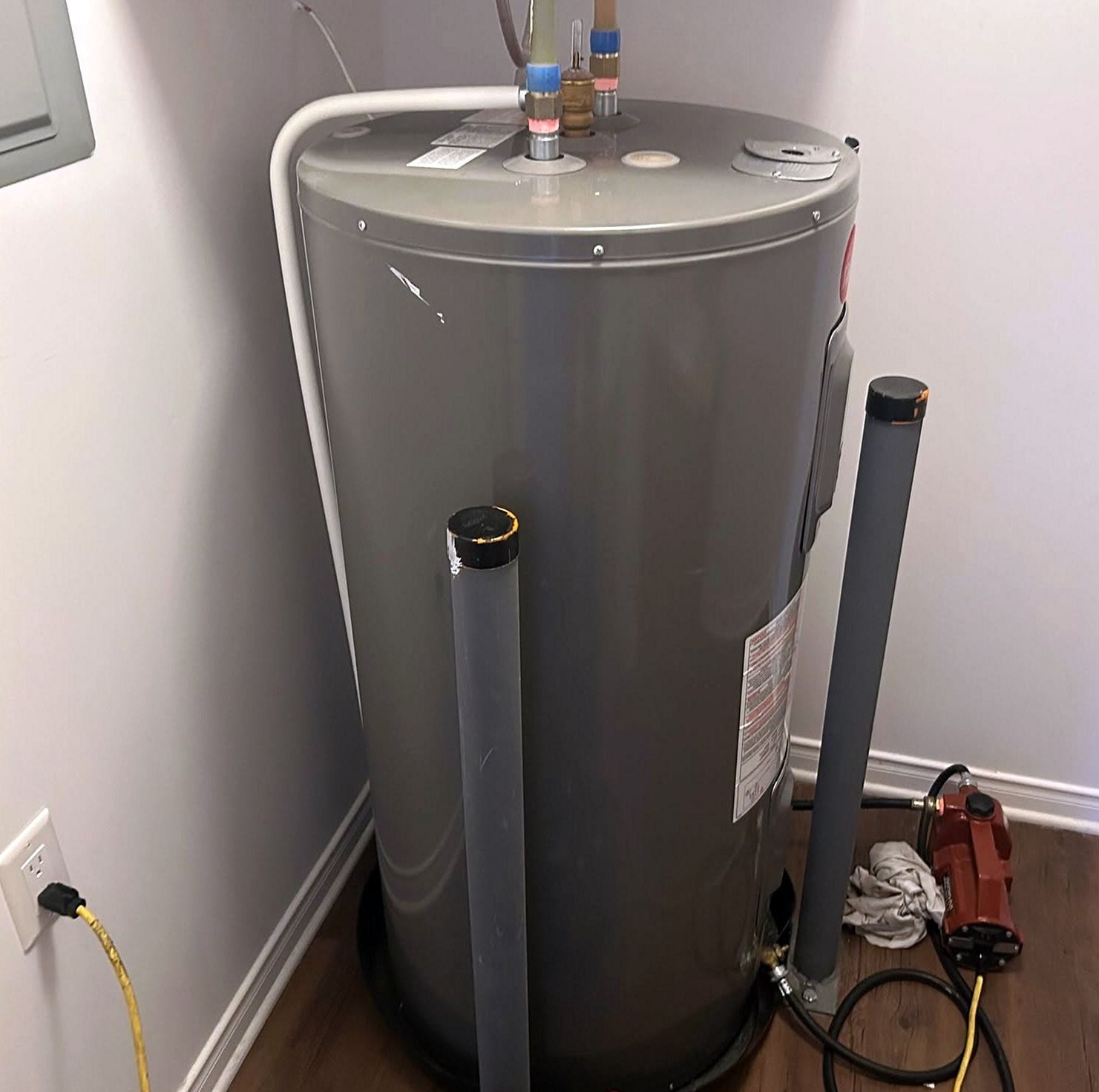 Gray electric tank water heater set in a utility closet, its supply lines and discharge pipes connected, tools still on the floor beside it