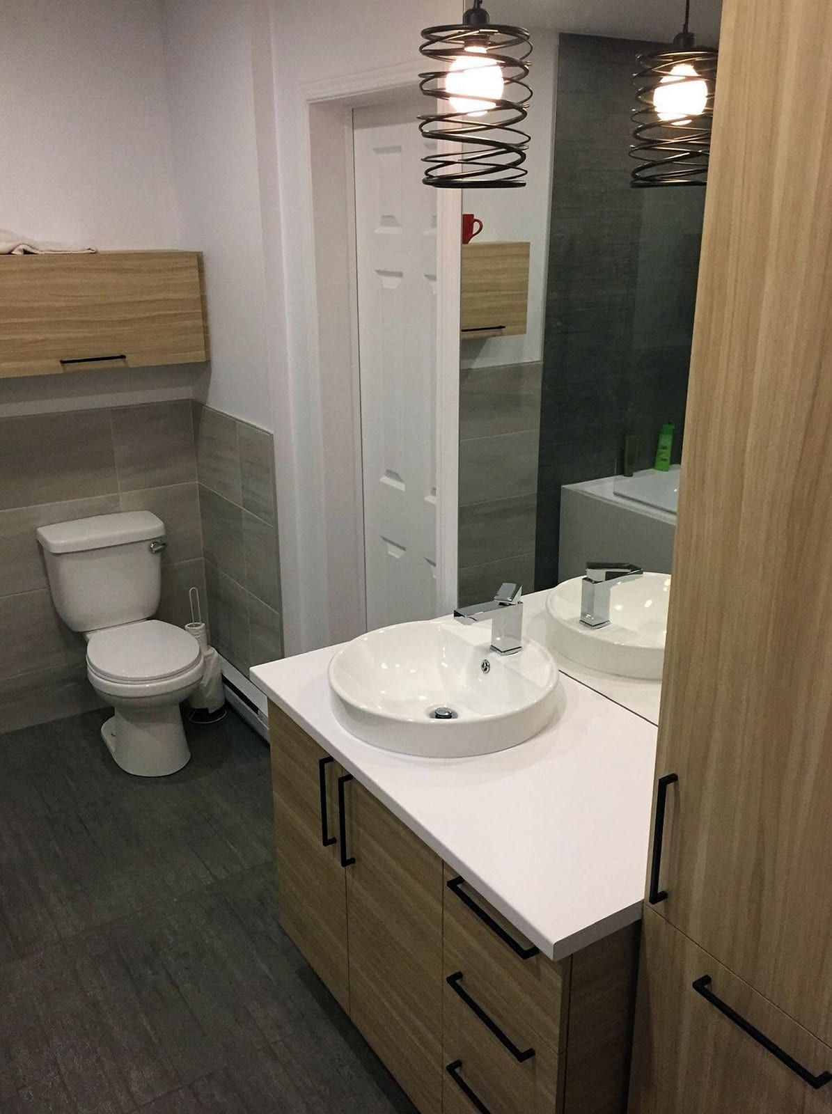 Bathroom with a round vessel sink on a light wood vanity, a toilet against gray tile and two pendant lights