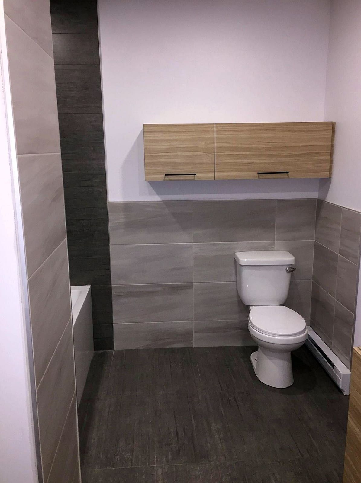 Toilet set against a wall of large gray tiles under a wall cabinet in a finished bathroom