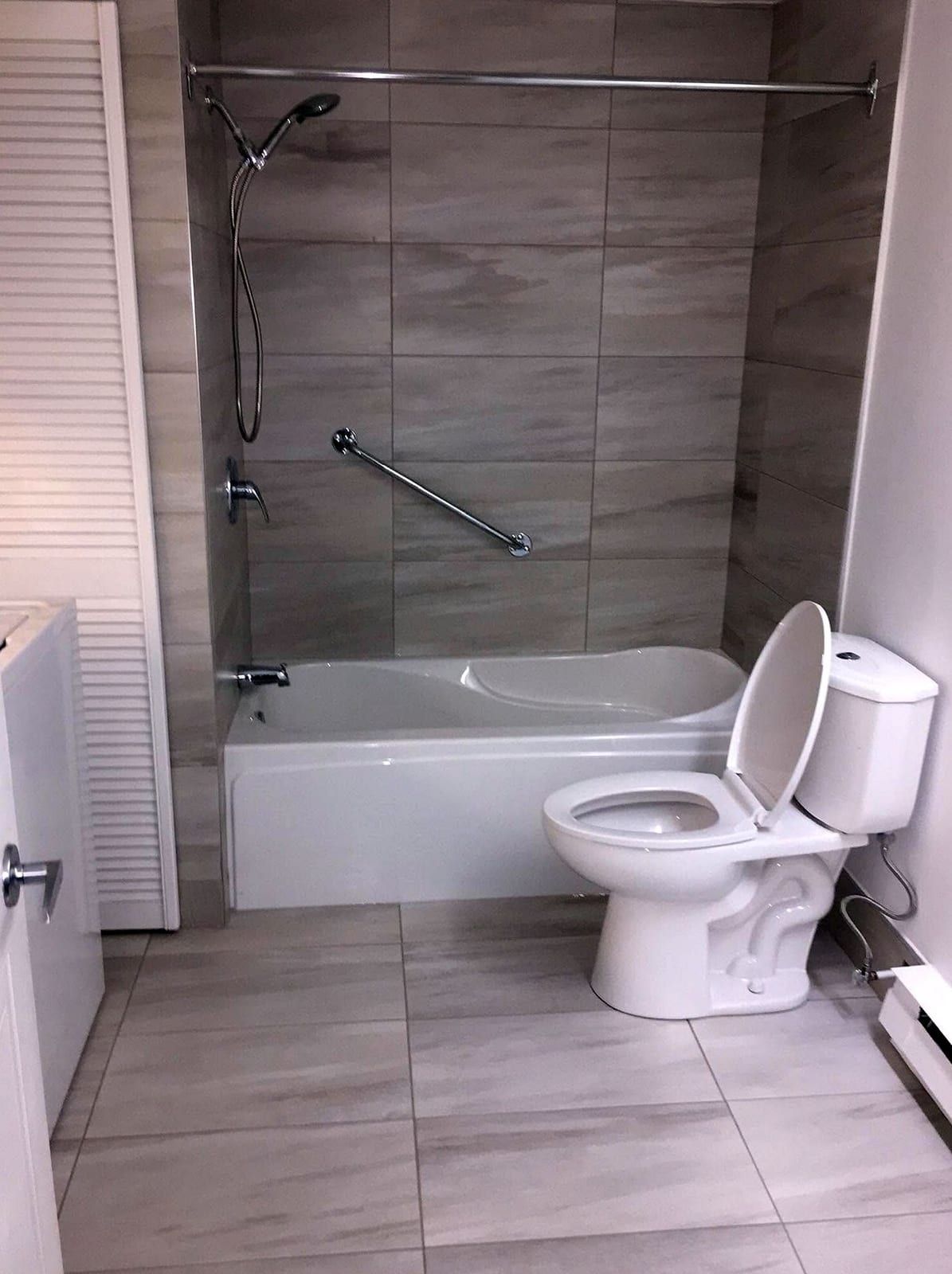 Bathtub with a handheld shower on a slide bar and a grab bar, a new toilet beside it on a tile floor