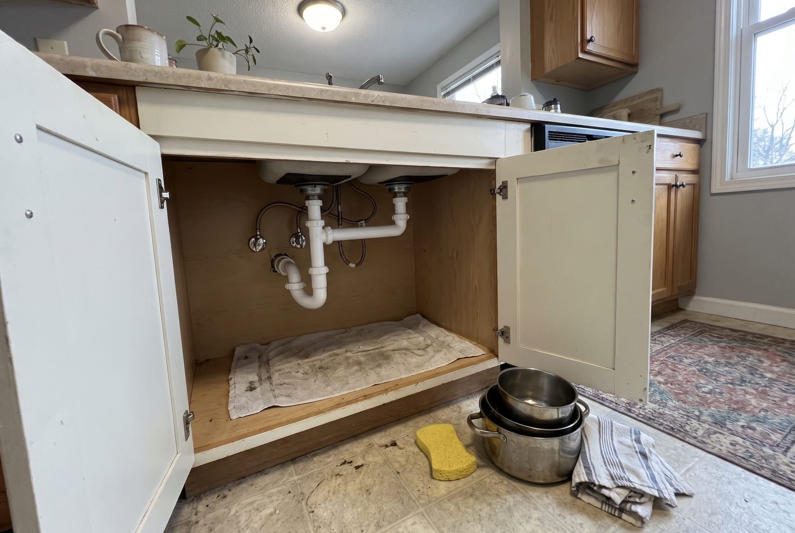 New white drain trap and chrome shut-off valves under a kitchen sink, the cabinet doors open and the pots set out on the floor