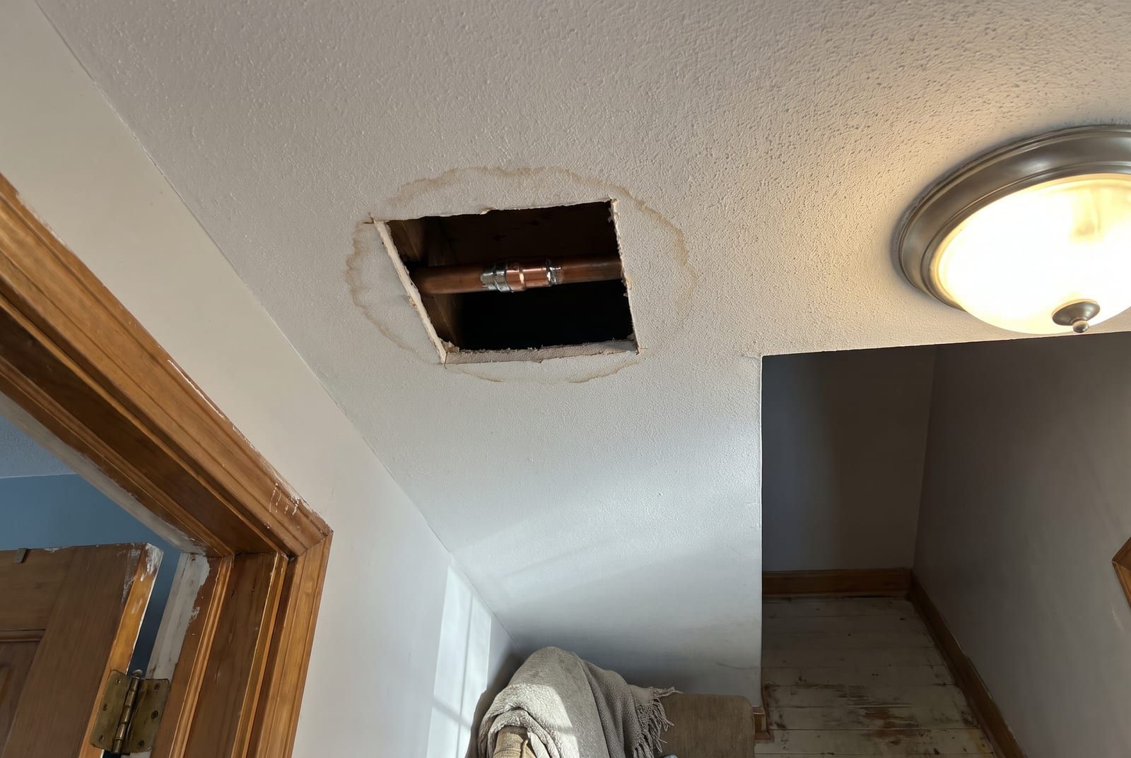 Square opening cut in a hallway ceiling, showing a copper water line with a repair coupling and a water stain around the opening