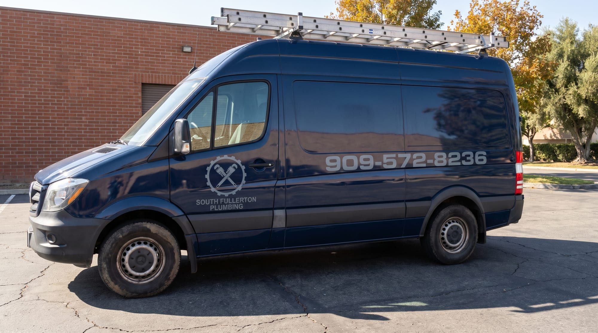 Navy Mercedes Sprinter work van with a ladder rack, the gear and crossed pipe wrench logo on the front door and the phone number on the cargo side, parked by a brick building